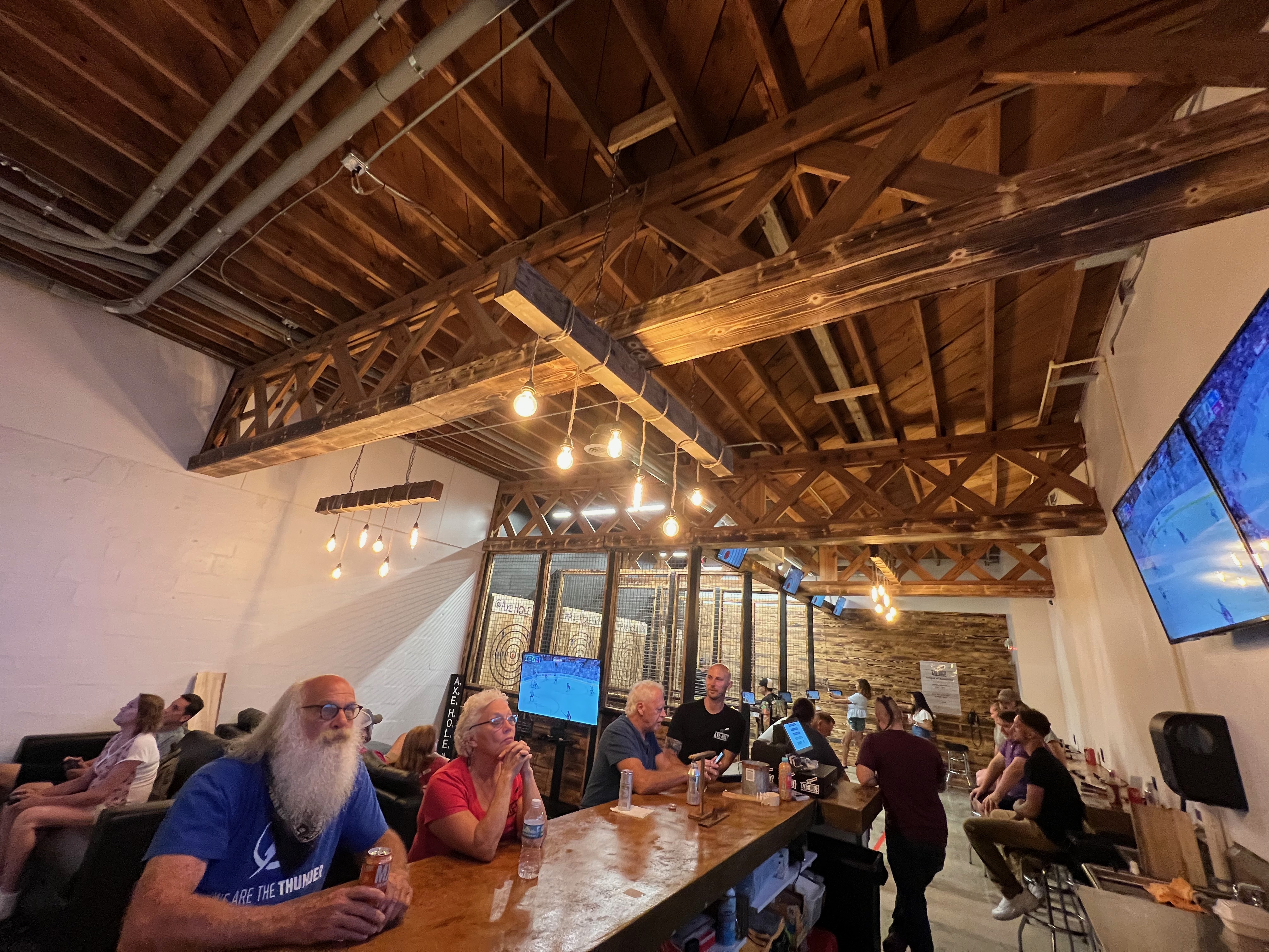 People sitting and enjoying drinks in a spacious room, with an axe throwing area visible in the background.